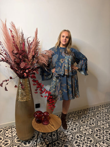 Een vrouw in een mat Bohemian kleed indigo staat met haar handen op haar heupen naast een hoge vaas met droogbloemen en een tafeltje met rood glazen decor, op tegels met een patroon tegen een effen muur.