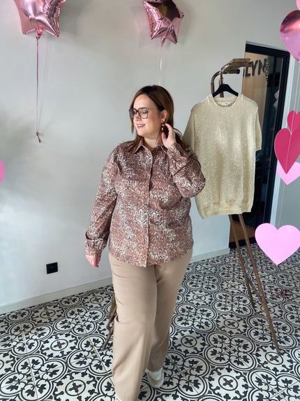 Vrouw met bril draagt een bruine slangenprint blouse gecombineerd met een beige wijde broek en witte sneakers, gefotografeerd in een  boutique waar ze kledij verkopen voor dames met een maatje extra 