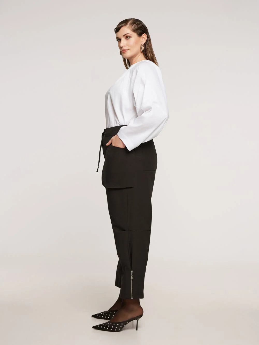Een vrouw staat in profiel tegen een effen achtergrond en draagt een witte blouse en de Zwarte Broek - Matmode met steekzakken voor een moderne look. Ze maakt haar outfit af met zwarte spitse hakken met witte stippen.