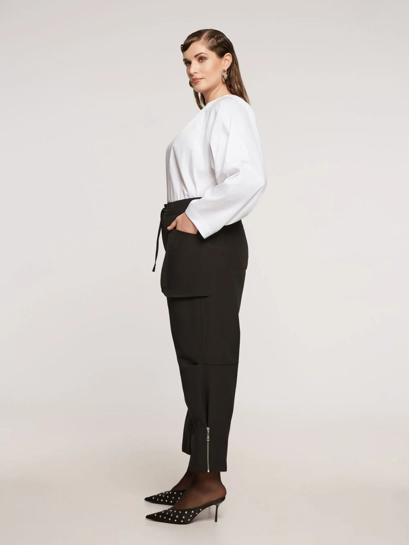 Een vrouw staat in profiel tegen een effen achtergrond en draagt een witte blouse en de Zwarte Broek - Matmode met steekzakken voor een moderne look. Ze maakt haar outfit af met zwarte spitse hakken met witte stippen.