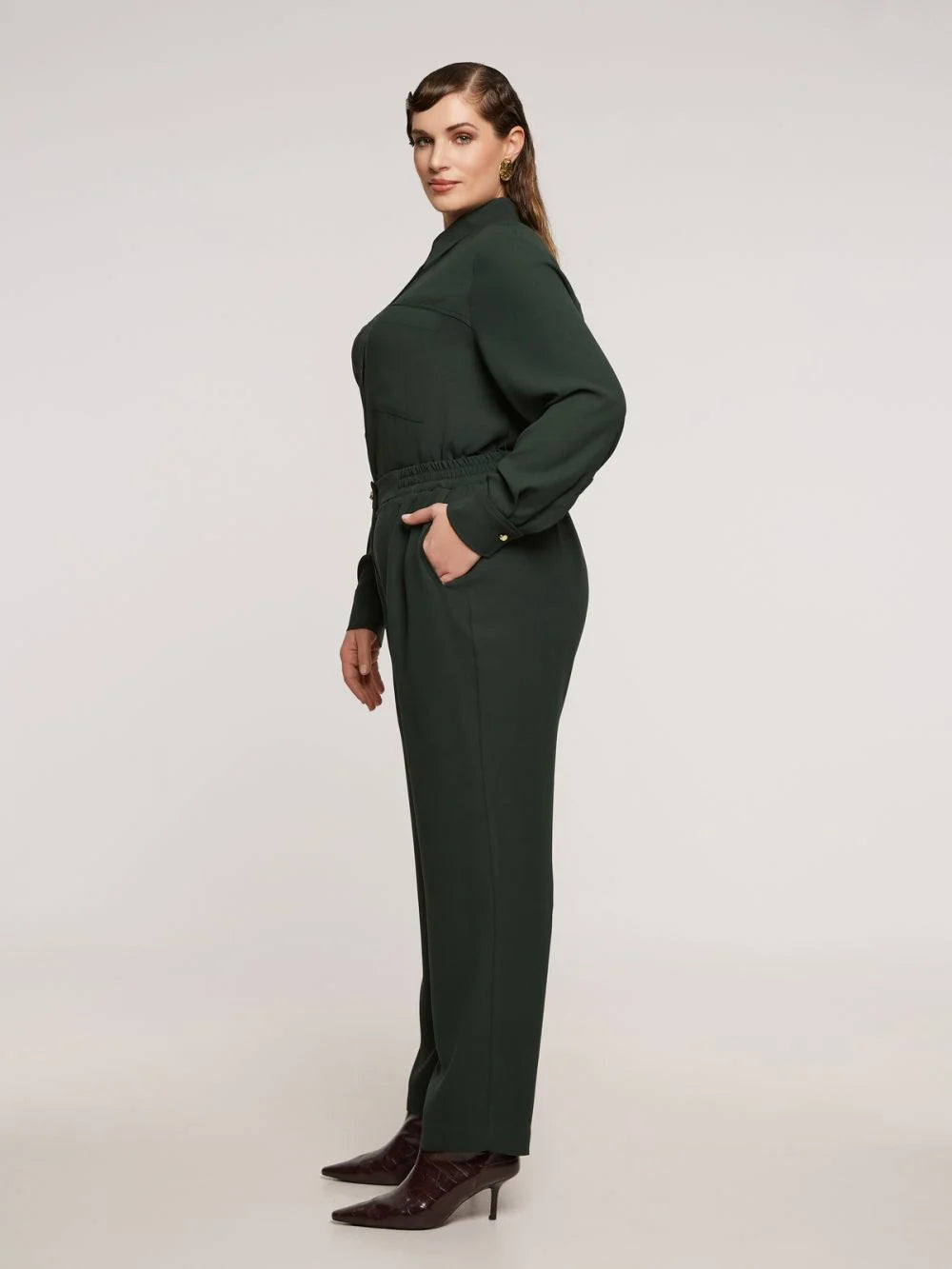Een vrouw met golvend bruin haar staat in profiel tegen een effen achtergrond, draagt een Groene broek - Mat Fashion, een donkergroene jumpsuit met lange mouwen van comfortabele, vloeiende stof en donkere laarzen met spitse tenen. Ze ziet er neutraal uit.