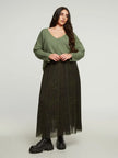 Een vrouw met lang donker haar en een neutrale uitdrukking staat tegen een effen achtergrond, draagt een losse olijfgroene trui, zwarte grove laarzen en de Donkergroene rok met print van Mat Fashion.
