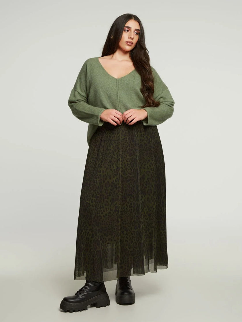 Een vrouw met lang donker haar en een neutrale uitdrukking staat tegen een effen achtergrond, draagt een losse olijfgroene trui, zwarte grove laarzen en de Donkergroene rok met print van Mat Fashion.