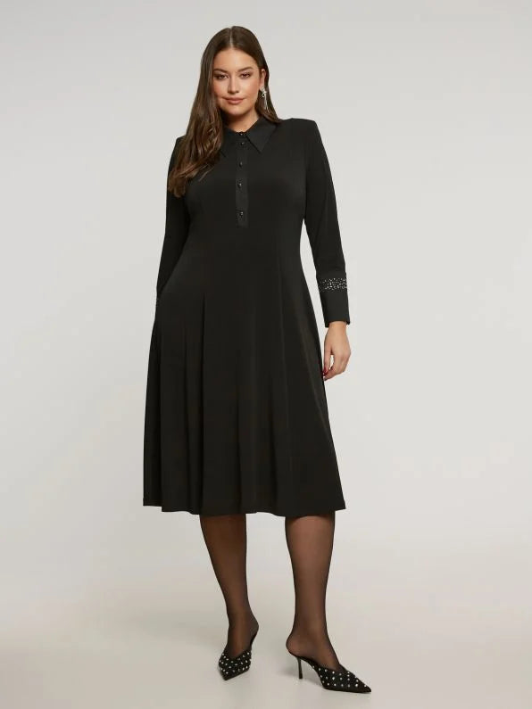 Een vrouw staat zelfverzekerd in het Zwart kleed van Mat Fashion - een zwarte jurk met lange mouwen, een kraag en knopen aan de voorkant. Ze draagt zwarte hakken met studs en heeft lang bruin haar.