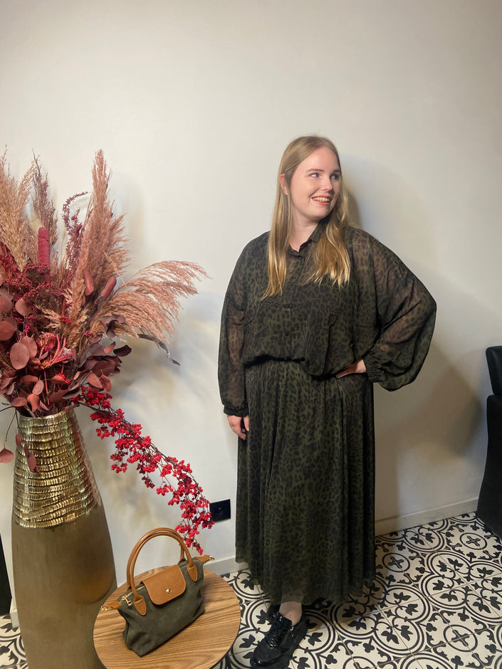 Een vrouw met lang blond haar draagt een blouse met Hemd met print - Mat Fashion en zwarte schoenen. Ze staat op vloertegels met patronen naast een hoge vaas met gedroogde bloemen. Een kleine groene handtas rust op een ronde houten tafel vlakbij.