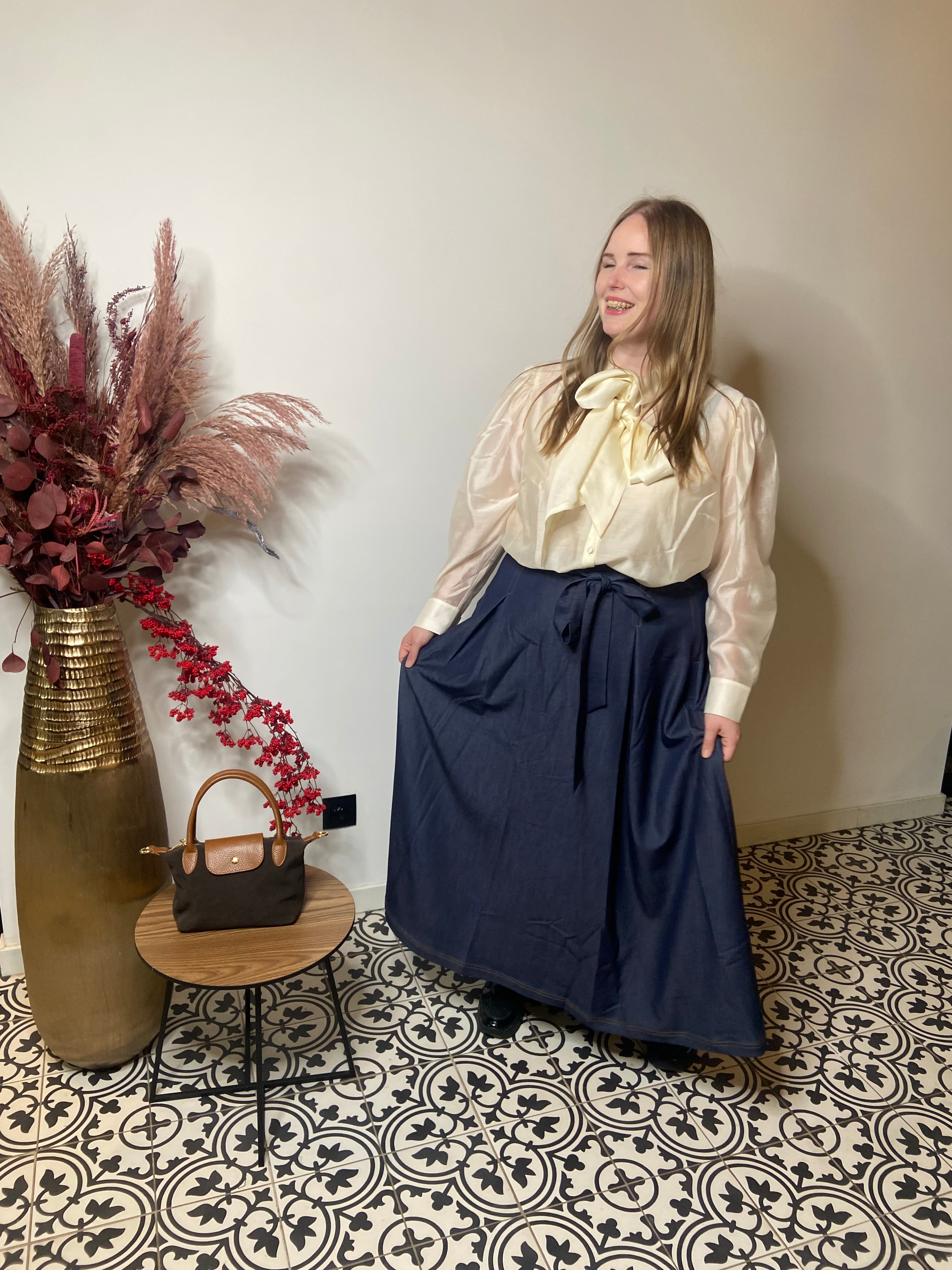 Een vrouw glimlacht in een kamer met gedessineerde tegels, ze draagt een crèmekleurige satijnen blouse met een strik en een trendy denim rok van Mat Fashion. Naast haar staat een tafeltje met een bruine tas en een hoge vaas gevuld met gedroogde bloemen.