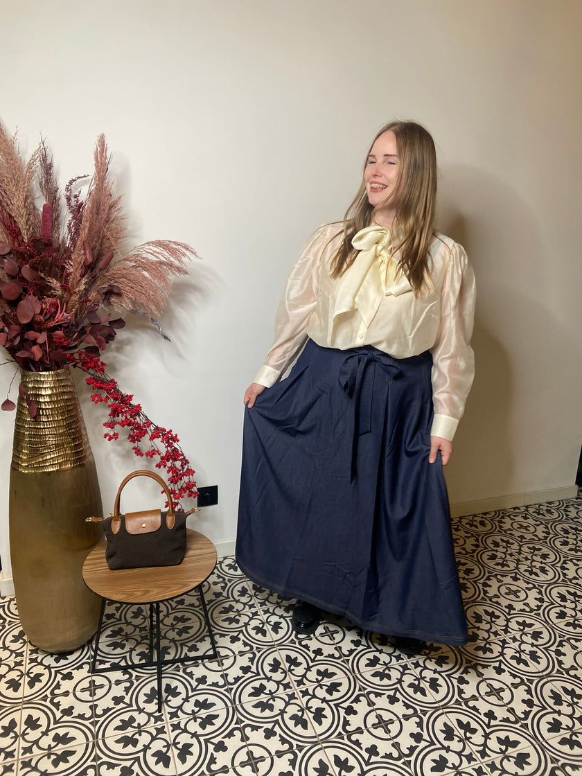 Een vrouw glimlacht in een kamer met gedessineerde tegels, ze draagt een crèmekleurige satijnen blouse met een strik en een trendy denim rok van Mat Fashion. Naast haar staat een tafeltje met een bruine tas en een hoge vaas gevuld met gedroogde bloemen.