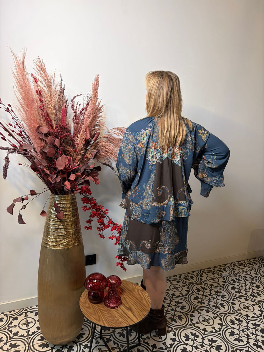 Een blonde vrouw staat met haar rug naar de camera en draagt de Bohemian kleed indigo van Mat Fashion. Ze staat naast een hoge vaas met gedroogde roze en rode planten, een tafeltje met rode glazen bollen en een zwart-wit gedessineerde vloer.