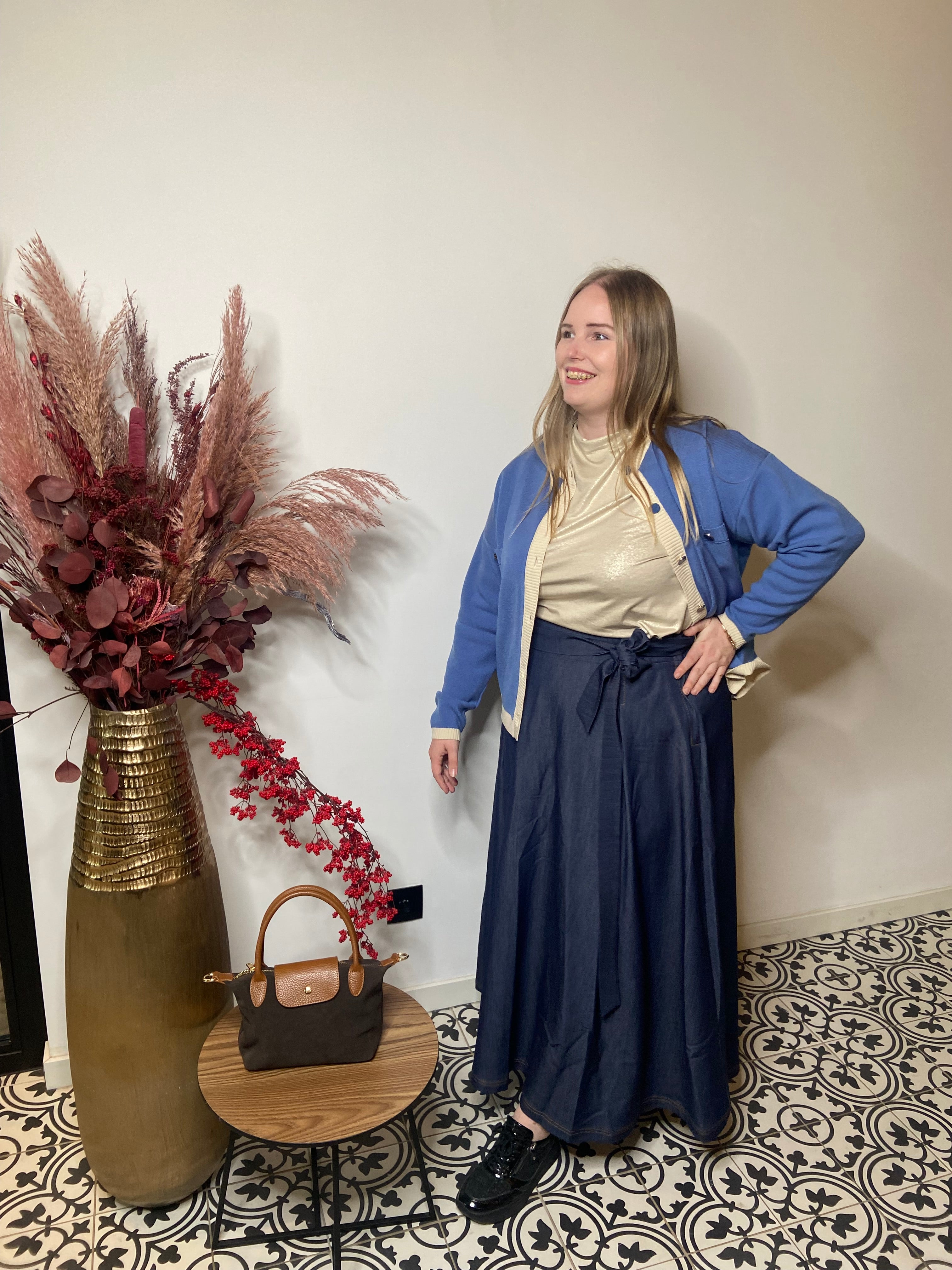 Een vrouw glimlacht binnenshuis op een tegelvloer met patroon. Ze draagt een lichte coltrui, een blauw vest en de Denim rok van Mat Fashion. Naast haar staan een vaas met gedroogde planten en een ronde tafel met een bruine handtas.