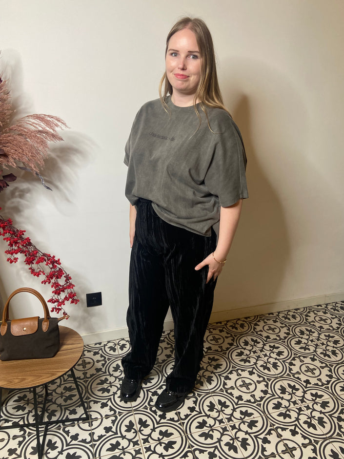 Een vrouw met lang blond haar staat binnen op tegels met een patroon, glimlachend in het T-shirt grijs - Mat Fashion, gecombineerd met een zwarte broek en schoenen. Een kleine bruine handtas rust op een nabijgelegen ronde tafel.