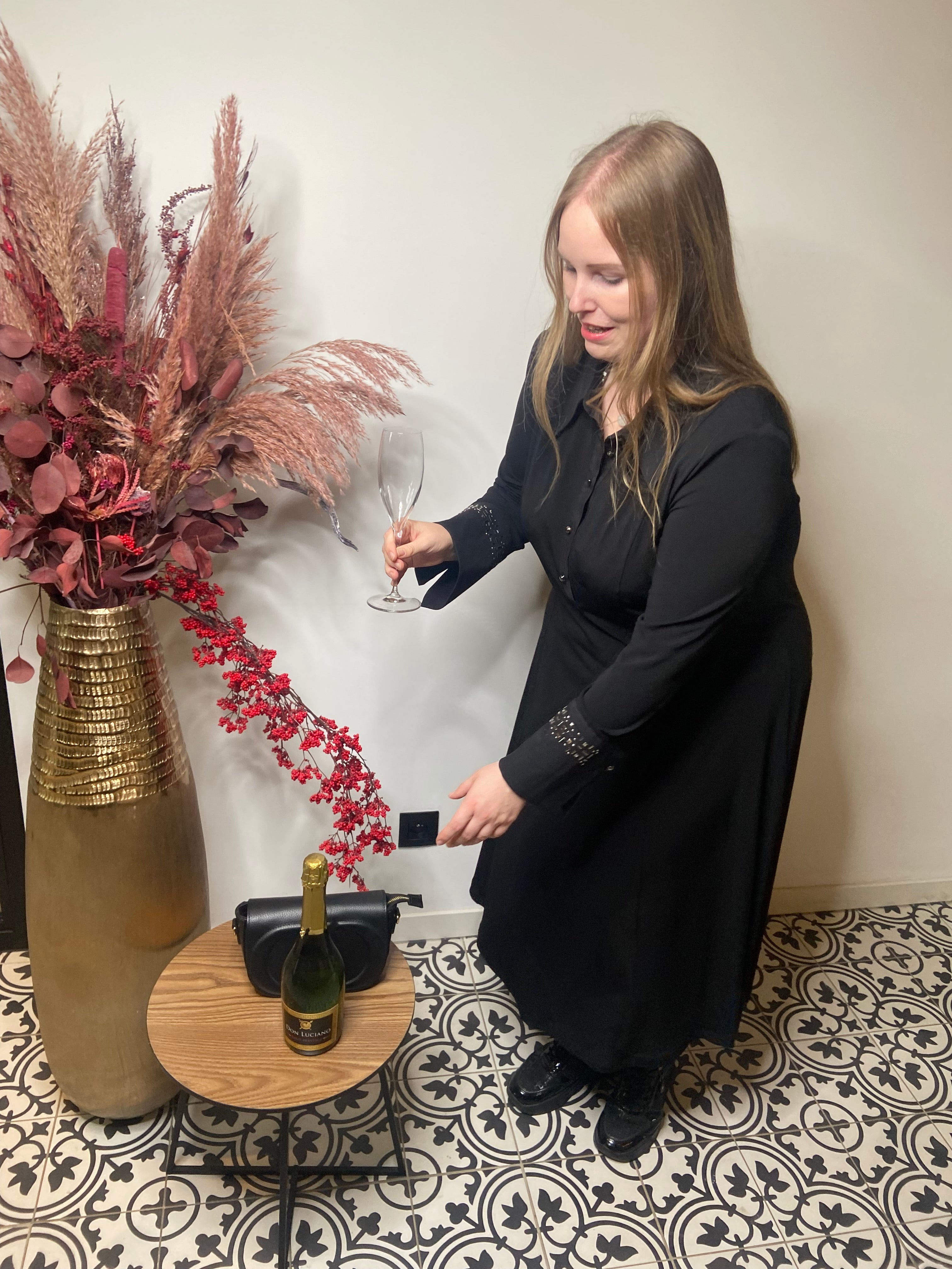 Een vrouw draagt het Zwart kleed van Mat Fashion en houdt een champagneglas vast bij een tafeltje met een fles champagne en een zwarte tas. Ze staat op patrontegels naast een hoge vaas met rode en roze gedroogde bloemen.