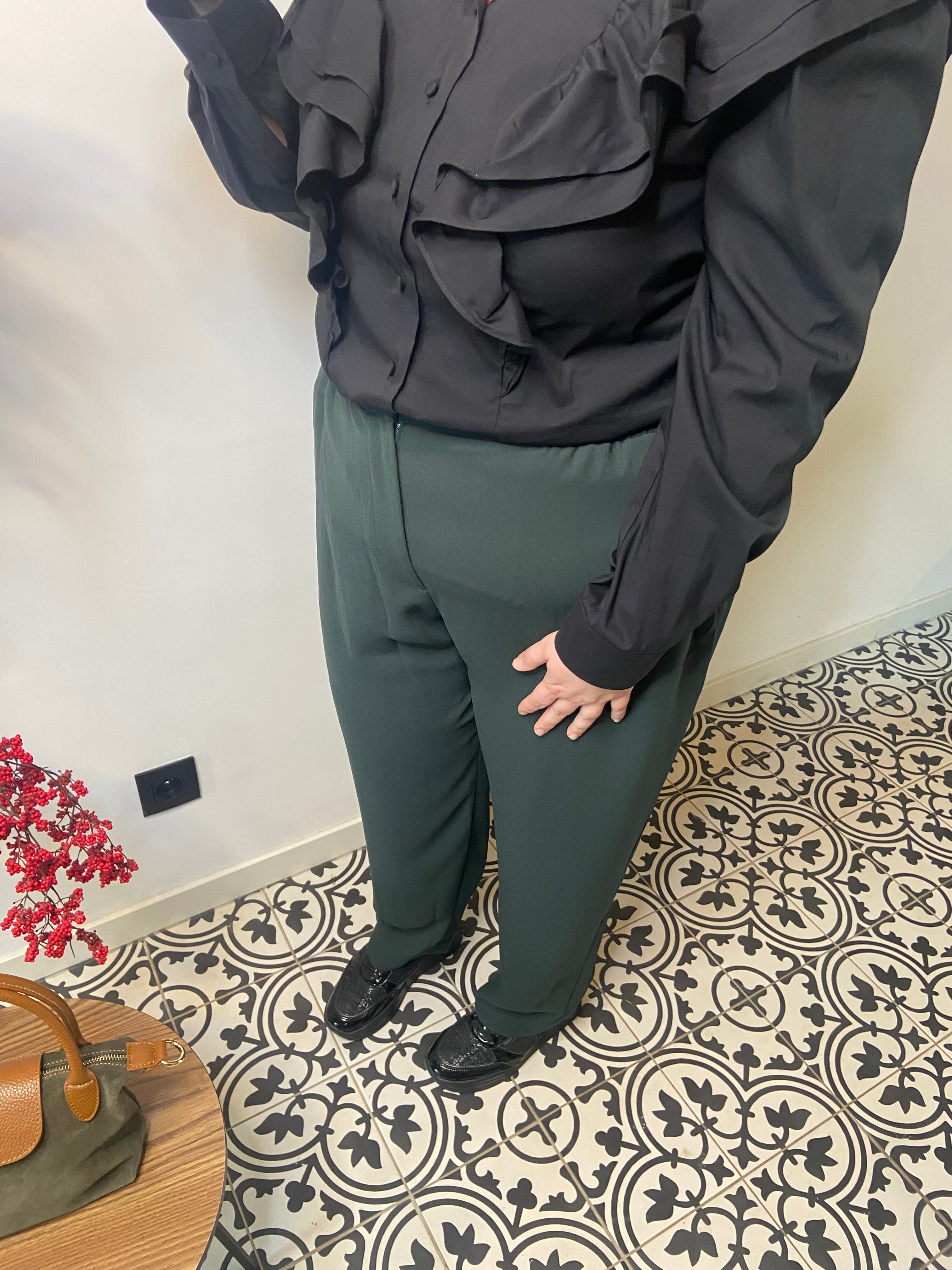 Een persoon staat op gedessineerde vloertegels, draagt een Groene broek - Mat Fashion, zwarte schoenen en een zwarte blouse met ruches. Een klein tafeltje met een tas en rode bloemen is zichtbaar in de linkerbenedenhoek.