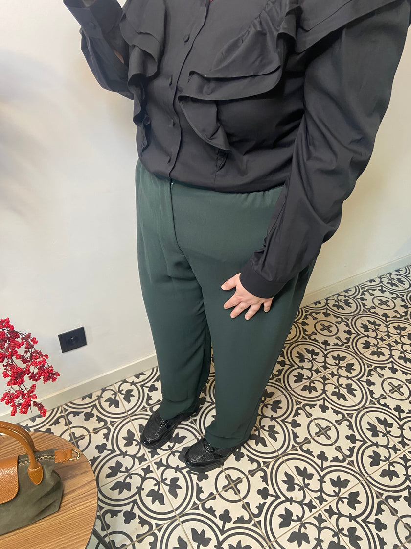 Een persoon staat op gedessineerde vloertegels, draagt een Groene broek - Mat Fashion, zwarte schoenen en een zwarte blouse met ruches. Een klein tafeltje met een tas en rode bloemen is zichtbaar in de linkerbenedenhoek.