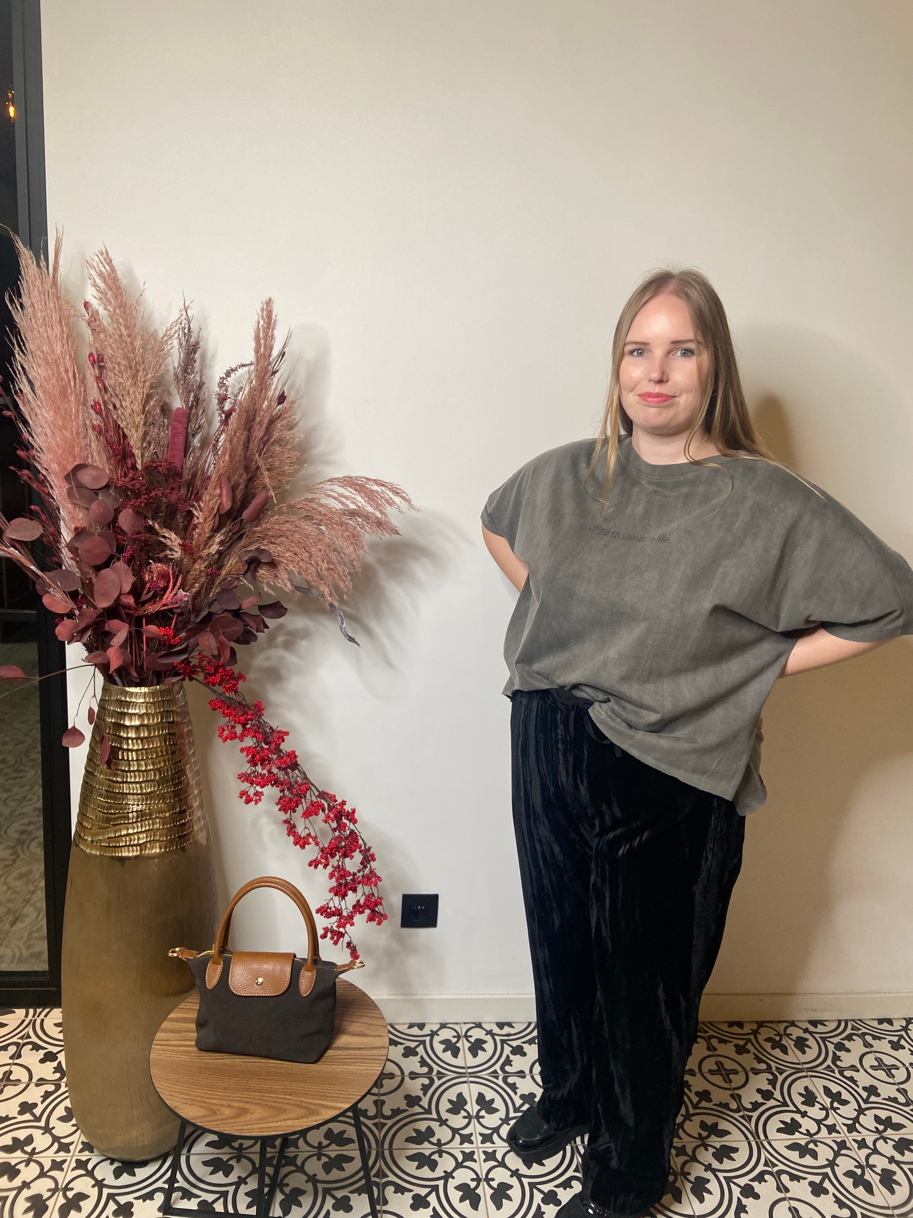 Een vrouw met steil blond haar staat binnen op tegels met een patroon, draagt het T-shirt grijs - Mat Fashion en een zwarte broek. Een gouden vaas met roze en bordeaux gedroogde bloemen versterkt de sfeer van Charismatic Life.