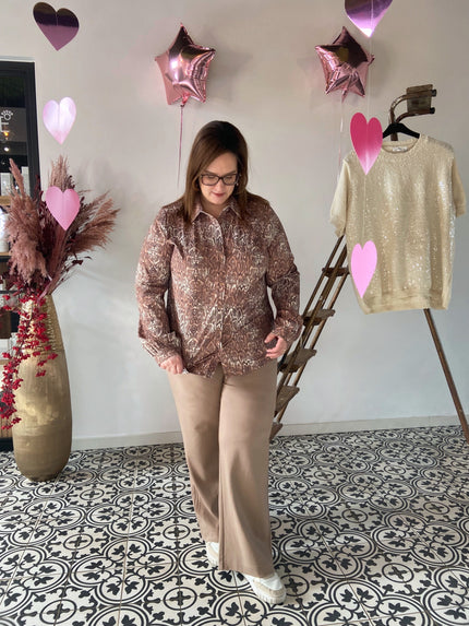 Vrouw met bril draagt een bruine slangenprint blouse gecombineerd met een beige wijde broek en witte sneakers, gefotografeerd in een boutique met decoratieve hart- en sterballonnen.
