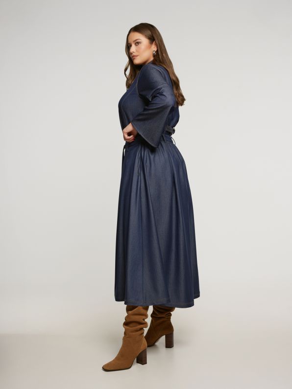 Een vrouw met lang bruin haar poseert zijdelings tegen een effen lichte achtergrond. Ze draagt de Denim rok van Mat Fashion - een lange, donkerblauwe jurk met wijde mouwen en een riem - in combinatie met bruine laarzen met hoge hakken voor een stijlvolle, rockgeïnspireerde look.