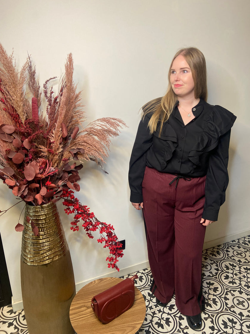 Een vrouw met lang blond haar staat binnen bij een vaas met gedroogde bloemen, draagt een zwarte blouse met ruches en een Rode broek met strepen - Mat Fashion, met een bijpassende handtas op een klein rond tafeltje naast haar.