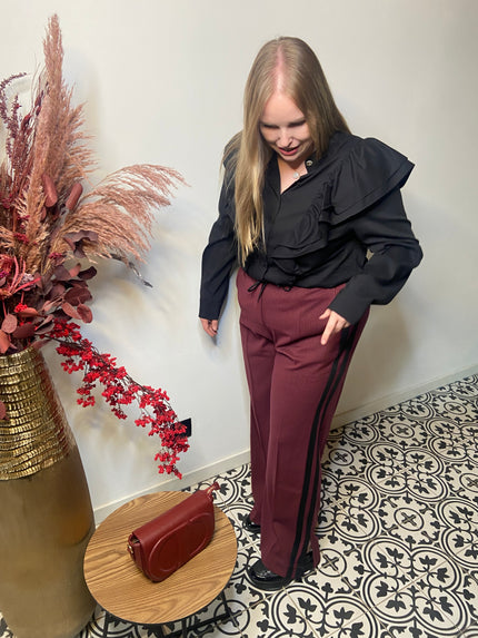 Een vrouw met lang blond haar in een zwarte blouse met ruches en een Rode broek met strepen van Mat Fashion staat op gedessineerde tegels bij een kleine ronde tafel met een rode tas en een hoge vaas met roze en rode droogbloemen voor een moderne sportieve look.