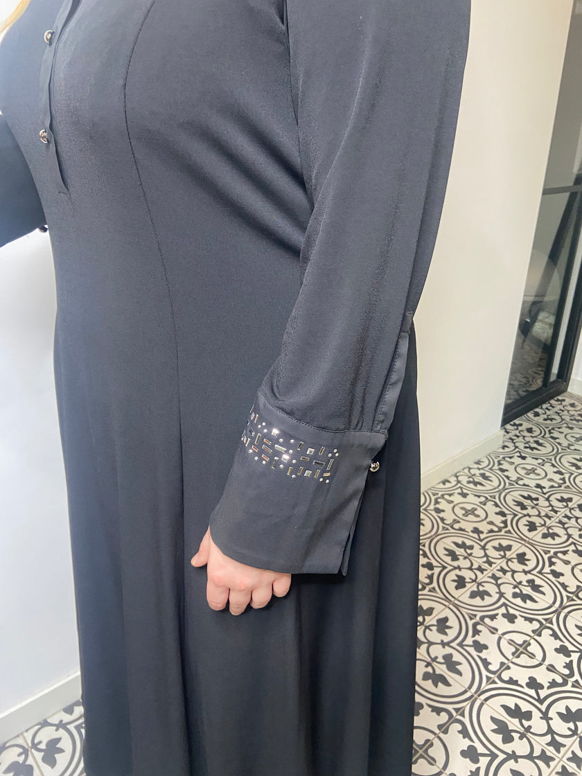 Een persoon met een Zwart kleed - Matte Mode met zilveren details op de mouwboorden staat op een tegelvloer met patroon bij een witte muur.