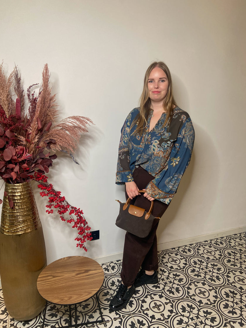 Een vrouw met lang blond haar staat binnen op gedessineerde vloertegels en draagt een Boheemse blouse van Mat Fashion met wijde mouwen, een donkere broek en zwarte schoenen. Ze houdt een bruine handtas vast naast een gouden vaas met gedroogde planten en een kleine ronde tafel.