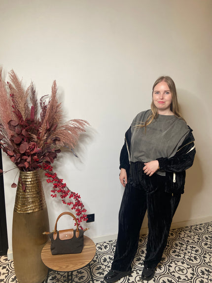 Een vrouw met lang blond haar staat binnen op tegels met een patroon en draagt het Zwarte jas huispak van Mat Fashion - een chique zwart polyester jasje over een grijs topje - naast een hoge vaas met gedroogde bloemen en een tafeltje met een bruine handtas.