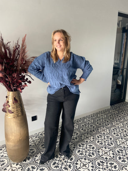 Een vrouw met blond haar glimlacht binnenshuis, draagt de Blauwe gebreide trui - Mat Fashion met een zwarte broek, staat op tegels met een patroon naast een hoge vaas met bordeauxrode gedroogde planten - een gezellige keuze voor elke wintergarderobe.