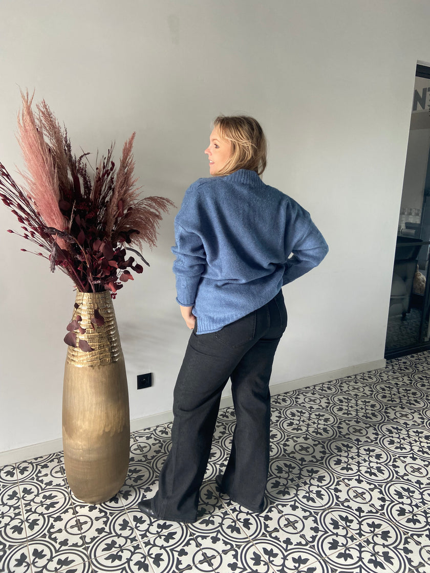 Een vrouw staat binnen op tegels met een patroon en draagt de Blauwe gebreide trui - Mat Fashion met een donkere broek. Naast haar staat een hoge gouden vaas gevuld met bordeauxrode gedroogde planten tegen een lichtgrijze muur.