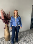 Een blonde vrouw staat binnen op een tegelvloer met patroon en draagt een blauw vest en de Jeansbroek - Mat Mode. Ze staat naast een grote gouden vaas met gedroogd bordeauxrood en beige gebladerte tegen een effen lichtgekleurde muur.