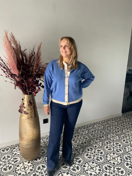 Een blonde vrouw glimlacht naast een hoge gouden vaas met gedroogd rood en roze gebladerte, draagt het Gebreide vest van Mat Fashion en een donkere spijkerbroek, op een tegelvloer met patroon tegen een witte muur. 61033300.