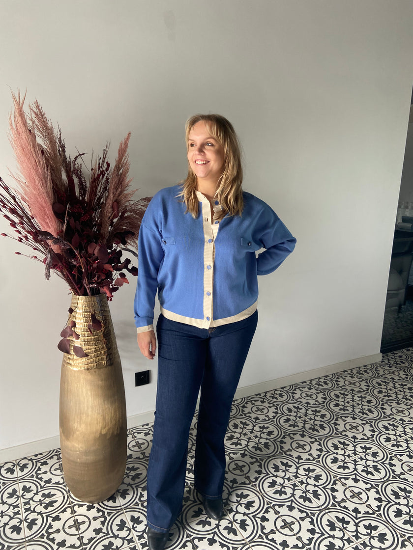 Een blonde vrouw glimlacht naast een hoge gouden vaas met gedroogd rood en roze gebladerte, draagt het Gebreide vest van Mat Fashion en een donkere spijkerbroek, op een tegelvloer met patroon tegen een witte muur. 61033300.