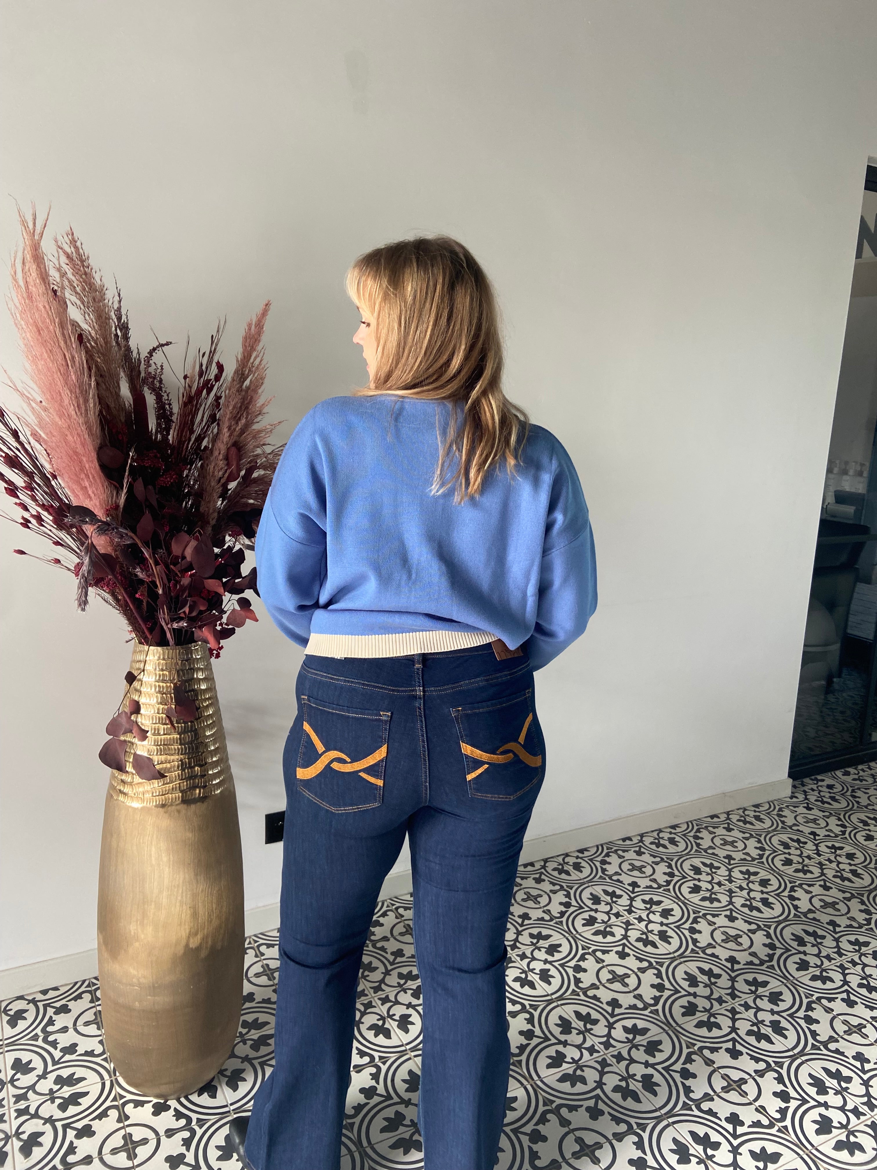Een vrouw staat binnen met haar rug naar de camera, draagt een blauwe trui en stijlvolle donkerblauwe Jeansbroek - Mat Mode met opgestikte zakmotieven, naast een hoge vaas met gedroogd roze en bruin gebladerte op een tegelvloer met patroon.