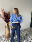 Een blonde vrouw in een lichtblauwe trui en Jeansbroek - Mat Fashion, met geelgestikte achterzakken, staat met haar gezicht naar een hoge gouden vaas met gedroogde bloemen op een tegelvloer met patroon.