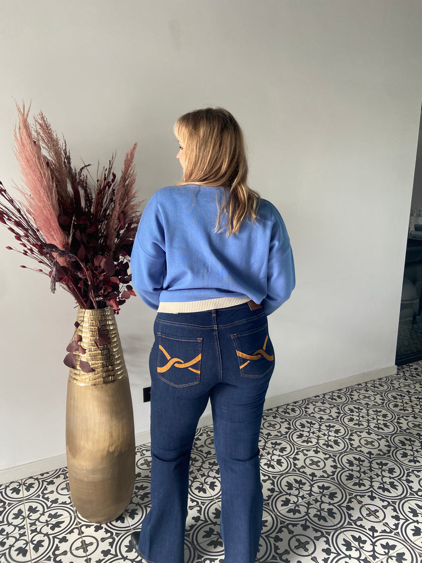 Een blonde vrouw in een lichtblauwe trui en Jeansbroek - Mat Fashion, met geelgestikte achterzakken, staat met haar gezicht naar een hoge gouden vaas met gedroogde bloemen op een tegelvloer met patroon.