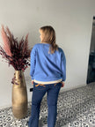 Een vrouw met blond haar, die het Gebreide denim vest van Mat Fashion en een donkere spijkerbroek draagt, staat met haar rug naar de camera op een tegelvloer met patroon naast een hoge gouden vaas gevuld met gedroogde planten.