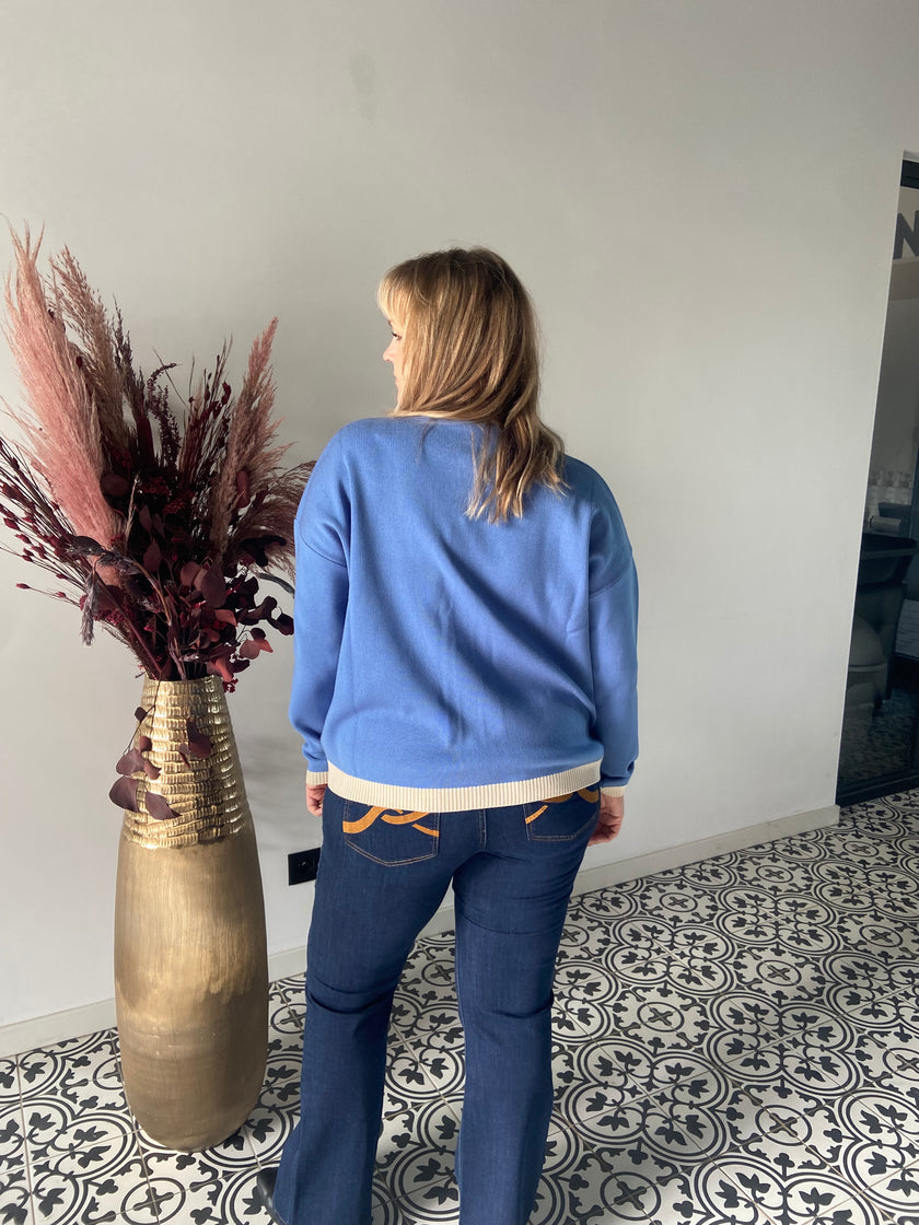 Een vrouw met blond haar, die het Gebreide denim vest van Mat Fashion en een donkere spijkerbroek draagt, staat met haar rug naar de camera op een tegelvloer met patroon naast een hoge gouden vaas gevuld met gedroogde planten.