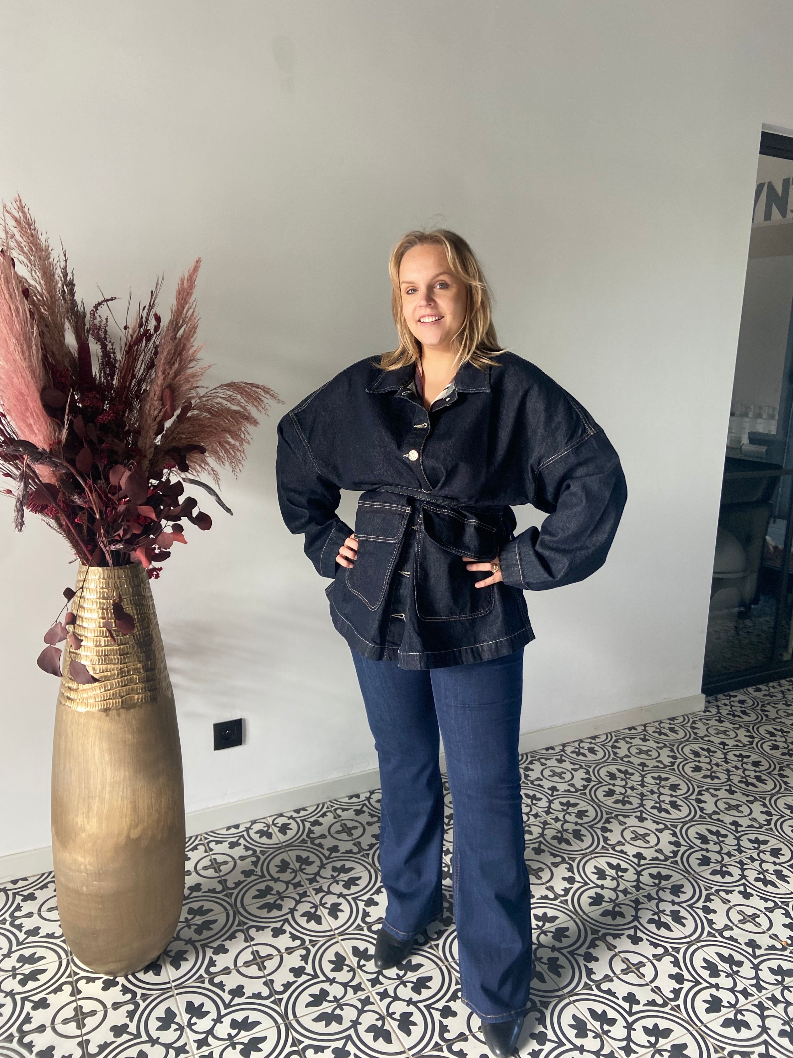 Een vrouw glimlacht in een stijlvolle kamer en draagt de Denim jas - Mat Fashion met blauwe jeans. Gedessineerde vloertegels en een hoge gouden vaas met gedroogde planten maken het plaatje compleet.