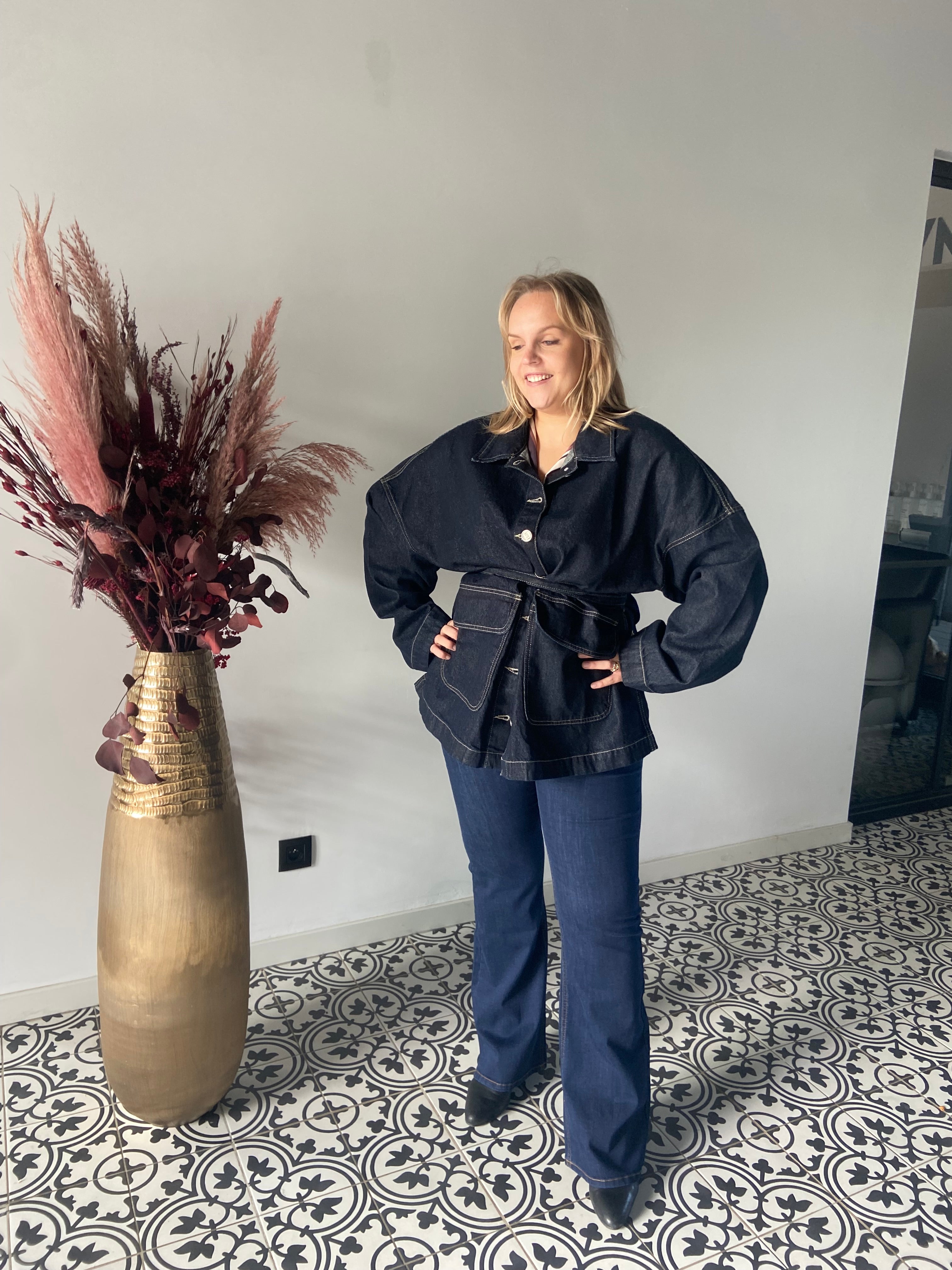 Een vrouw glimlacht binnenshuis op een tegelvloer met patroon. Ze draagt de Denim jas van Mat Fashion die in de taille is vastgebonden met een donkere spijkerbroek. Ze staat naast een hoge gouden vaas gevuld met gedroogd pampagras en bordeauxrood gebladerte.