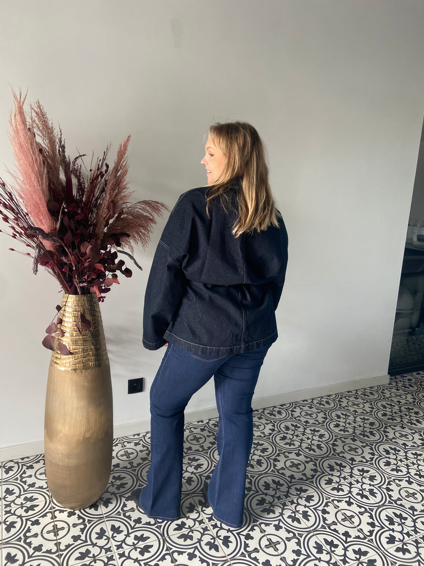Een blonde vrouw, die de Denim jas - Mat Fashion en flared jeans draagt, staat binnen op gedessineerde tegels met haar gezicht weg van een hoge gouden vaas met roze en bordeauxrode droogbloemen tegen een lichtgrijze muur.