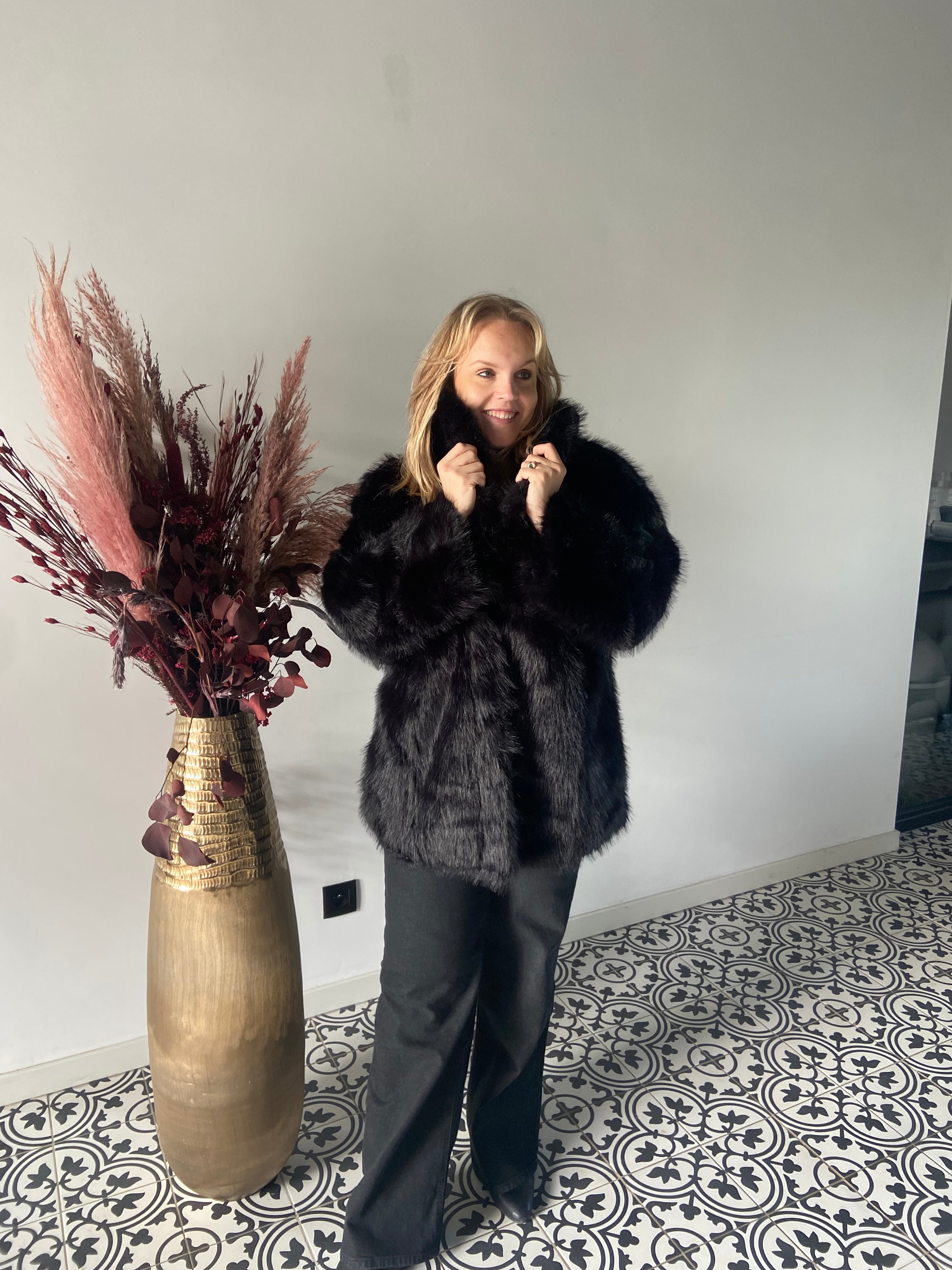 Een blonde vrouw glimlacht terwijl ze een Zwarte bontjas van Mat Fashion draagt. Ze staat binnen op een tegelvloer met patroon naast een hoge gouden vaas gevuld met gedroogde planten.