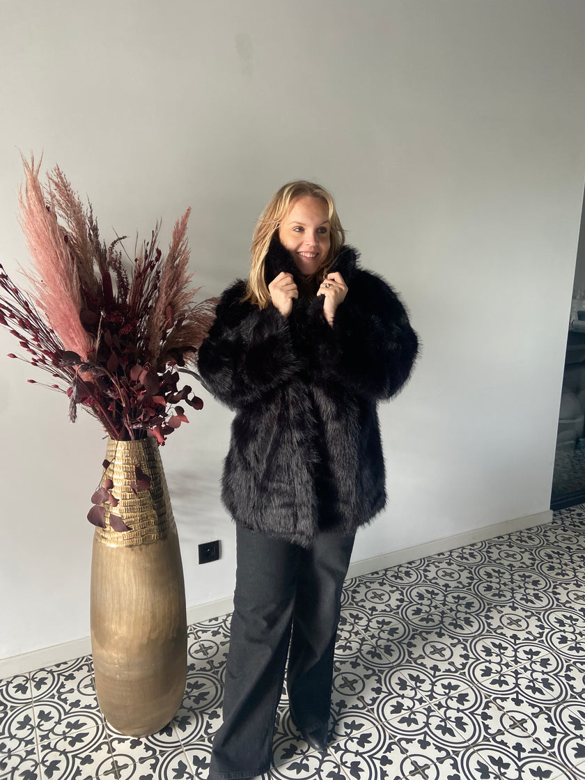 Een blonde vrouw glimlacht terwijl ze een Zwarte bontjas van Mat Fashion draagt. Ze staat binnen op een tegelvloer met patroon naast een hoge gouden vaas gevuld met gedroogde planten.