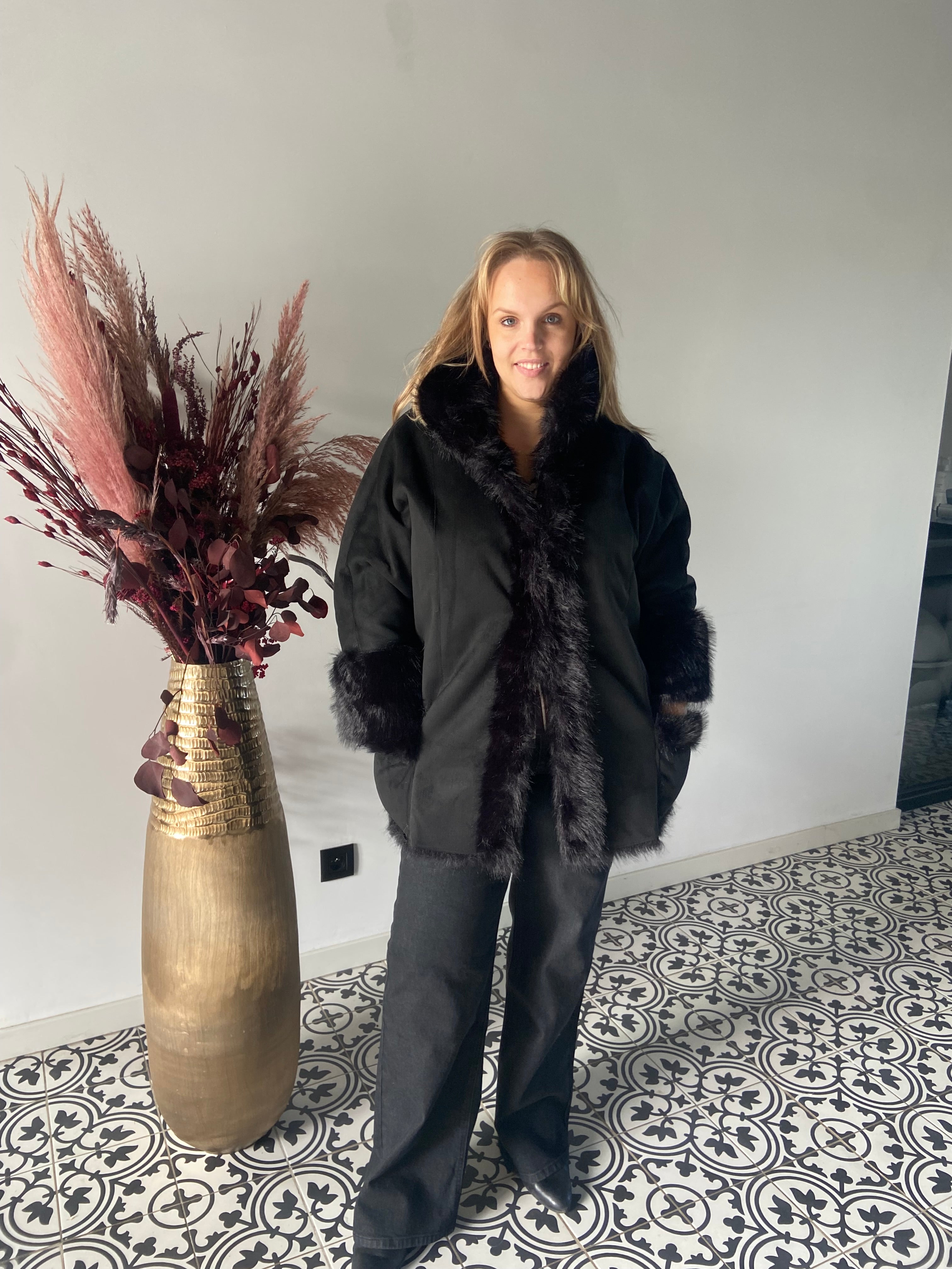 Een vrouw in een Mat Fashion Zwarte bontjas en zwarte broek staat binnen op een tegelvloer met patroon naast een hoge gouden vaas met gedroogde bordeauxrode en beige planten, tegen een effen lichte muur.