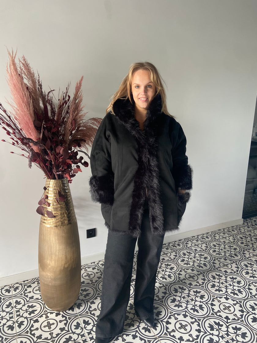 Een vrouw in een Mat Fashion Zwarte bontjas en zwarte broek staat binnen op een tegelvloer met patroon naast een hoge gouden vaas met gedroogde bordeauxrode en beige planten, tegen een effen lichte muur.