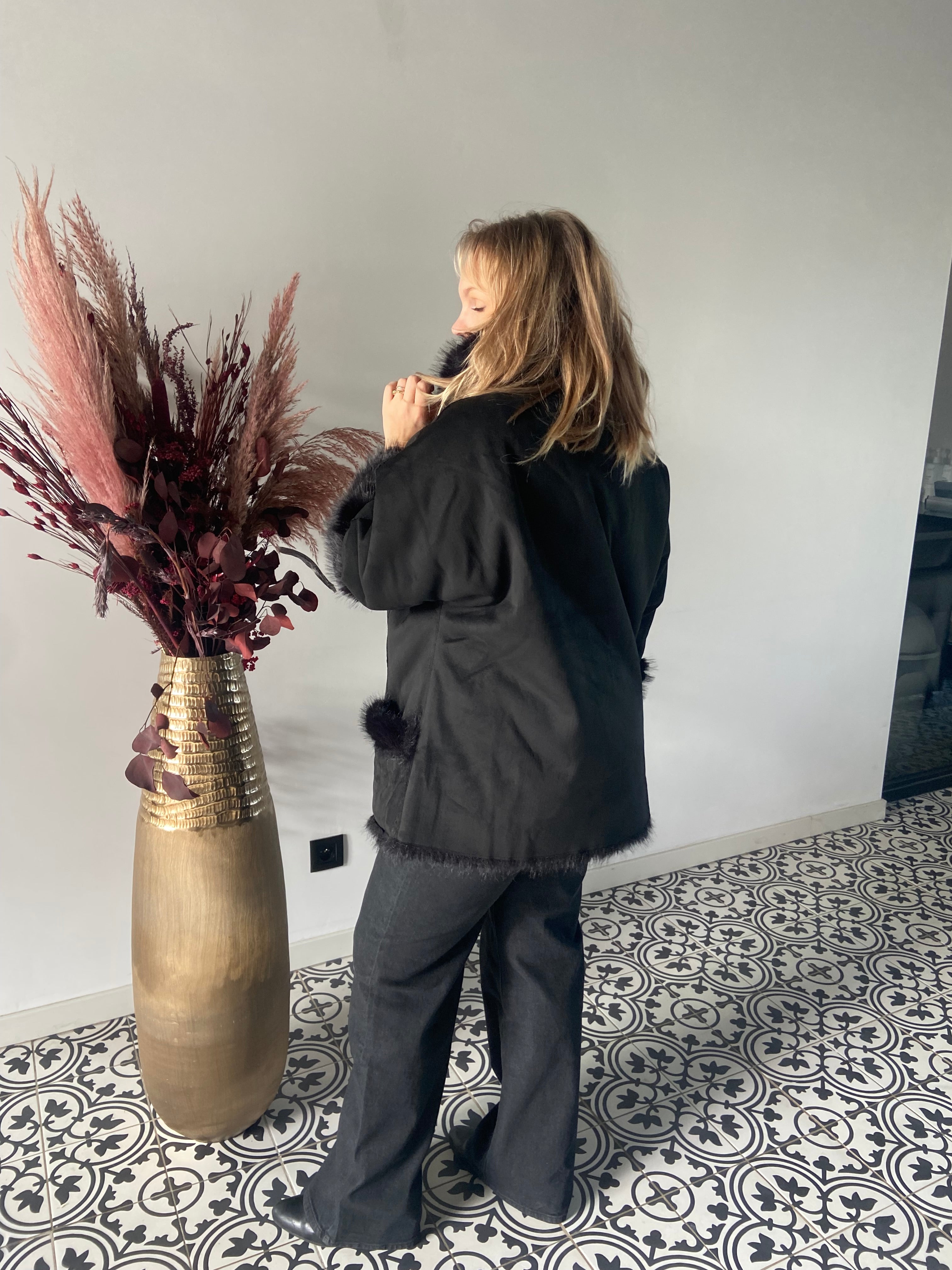 Een blonde vrouw staat binnen op een tegelvloer met patroon, draagt de Zwarte bontjas van imitatiebont - Matmode en een donkere broek, met haar gezicht van de camera afgewend naast een hoge koperen vaas gevuld met roze en bordeauxrode gedroogde planten.