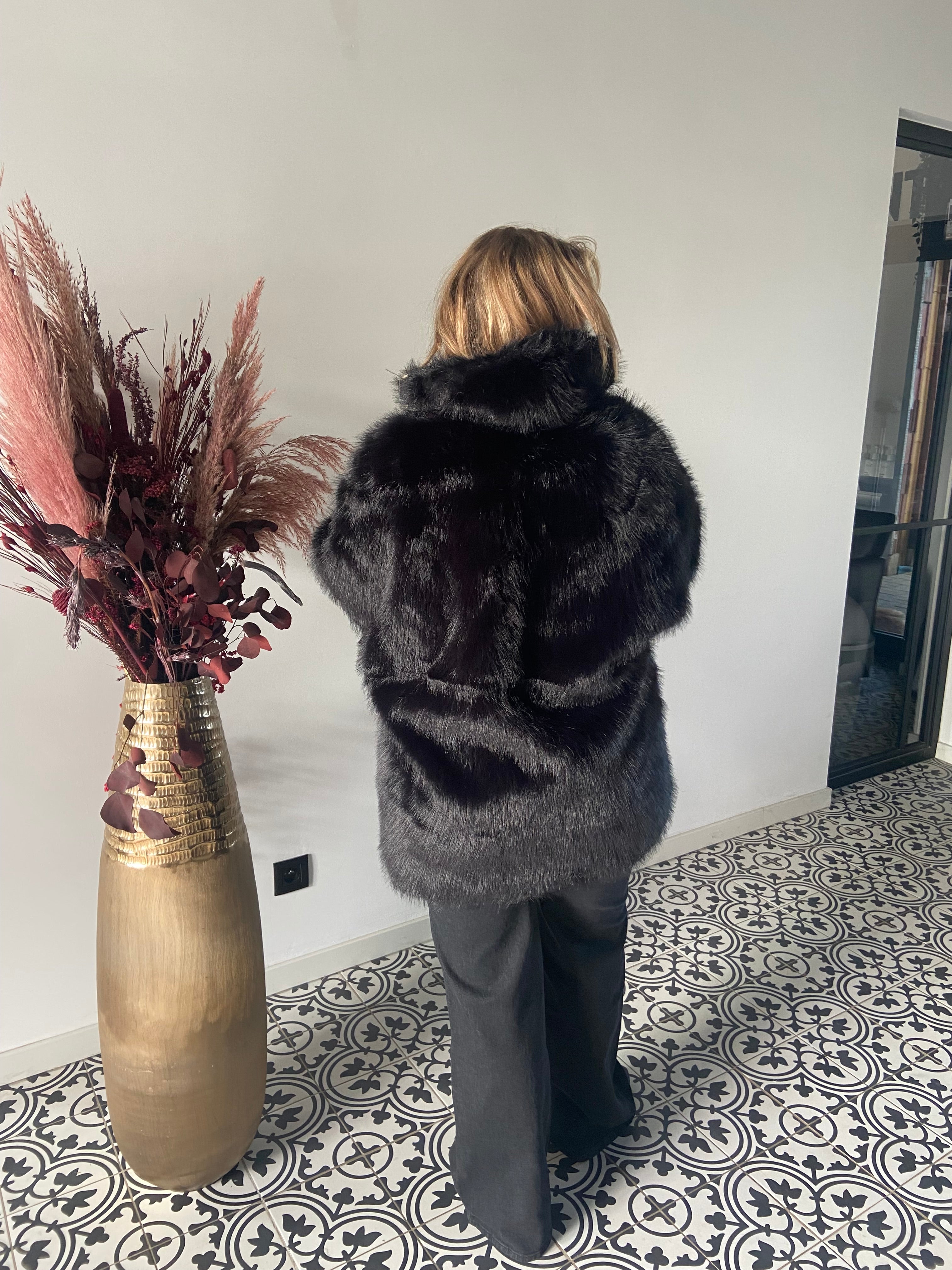 Een persoon met lichtbruin haar, van achteren gezien, draagt een Zwarte bontjas van imitatiebont en een donkere broek. Hij staat op een tegelvloer met patroon naast een hoge vaas gevuld met gedroogde planten.