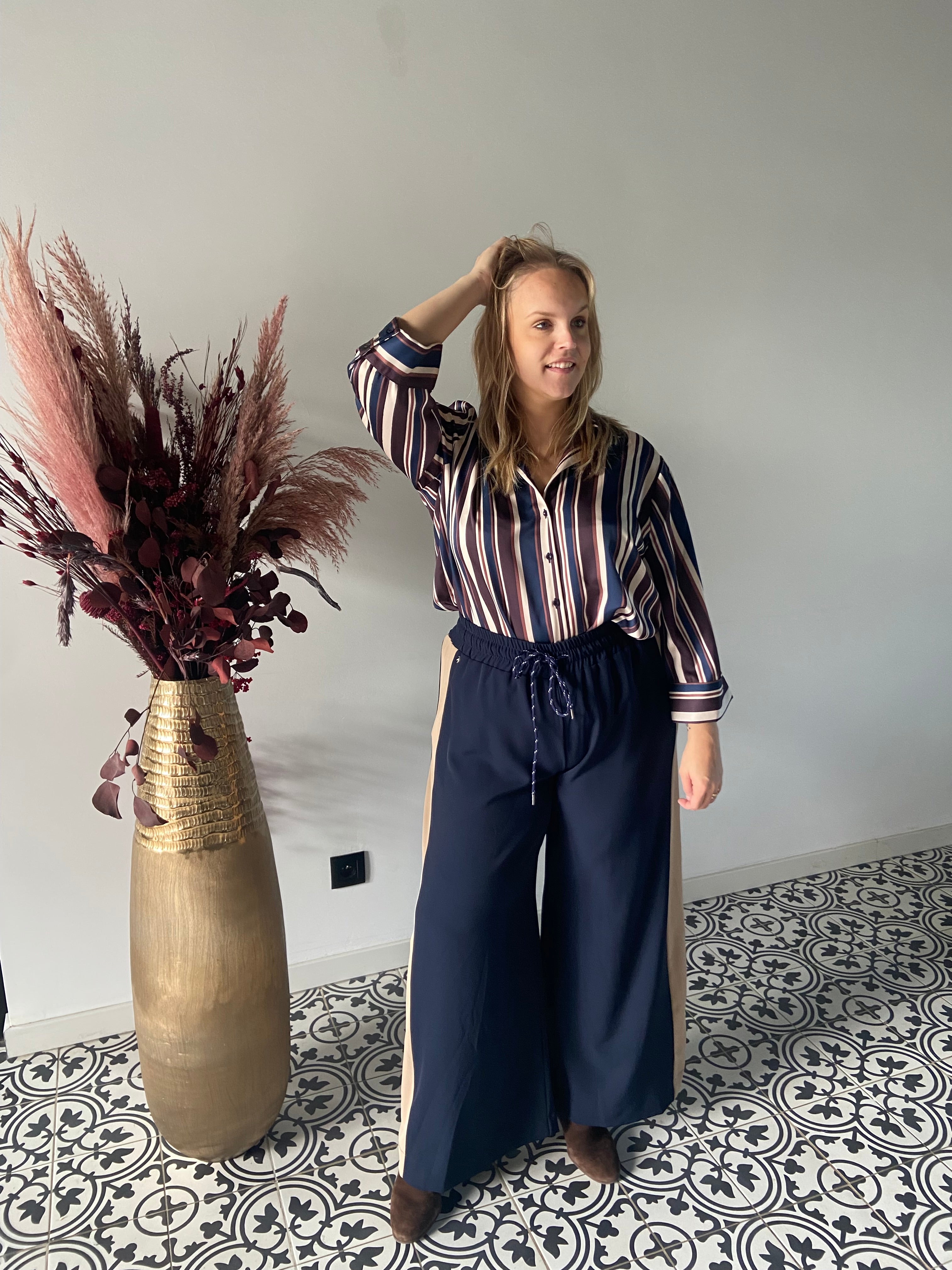 Een vrouw staat binnen op tegels met een patroon, draagt de Epicea hemd in marineblauw en een broek met wijde pijpen. Ze glimlacht lichtjes, een hand raakt haar hoofd. Een hoge gouden vaas met gedroogd roze en mauve gebladerte staat naast haar tegen een effen muur.