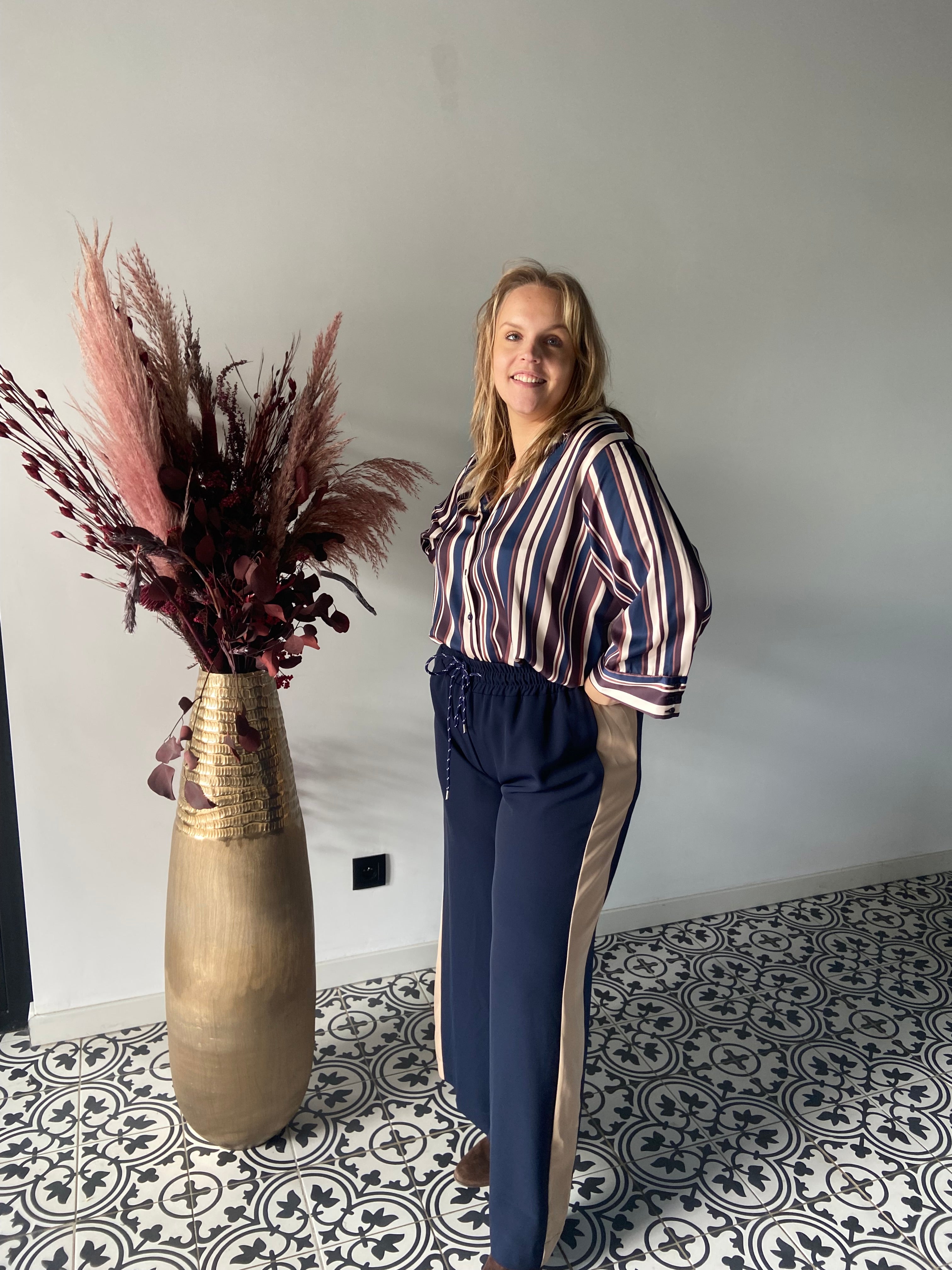 Een vrouw glimlacht naar de camera binnenshuis op een tegelvloer met patroon, draagt de Epicea hemd - Navy blouse en navy broek met tan zijstrepen, staande naast een hoge gouden vaas gevuld met gedroogd rood en roze gebladerte.