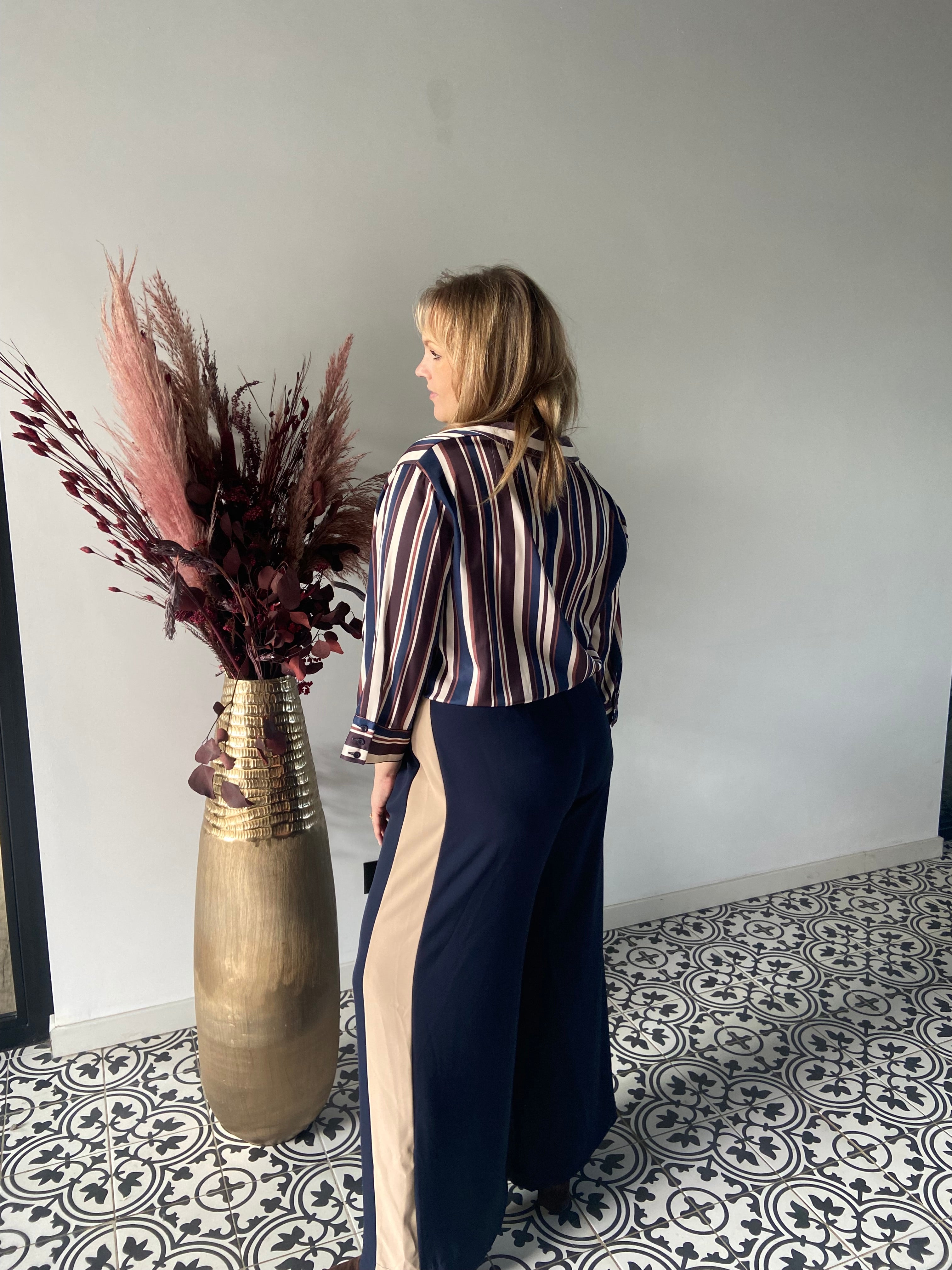 Een vrouw met blond haar, die de Epicea hemd - Navy en een wijde marine broek met beige panelen draagt, staat met haar rug naar de camera bij een hoge gouden vaas gevuld met gedroogd bordeauxrood en roze gebladerte. Gedessineerde vloertegels zijn zichtbaar.