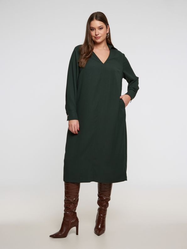 Een vrouw poseert zelfverzekerd in de Groen kleed - Mat mode, een donkergroene, knielange jurk met lange mouwen en een V-hals, gestyled met bruine kniehoge laarzen tegen een effen witte achtergrond.
