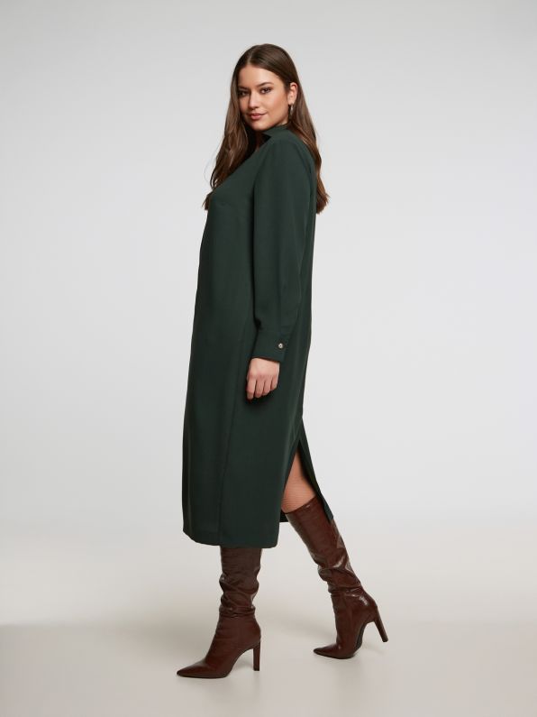 Een vrouw staat opzij in een Groen kleed - Matmode met lange mouwen en een split op de rug, gecombineerd met bruine kniehoge hakken. De achtergrond is effen en lichtgekleurd.