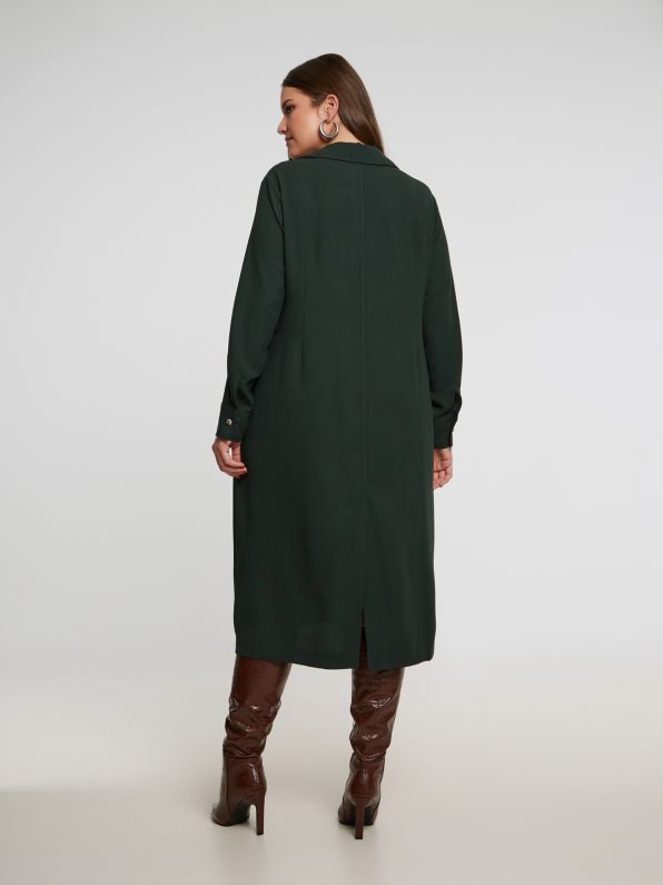 Een vrouw staat met haar gezicht naar voren in een Groen kleed - Mat Fashion, gecombineerd met bruine kniehoge laarzen, moeiteloos stijlvol tegen een lichte studio-achtergrond.